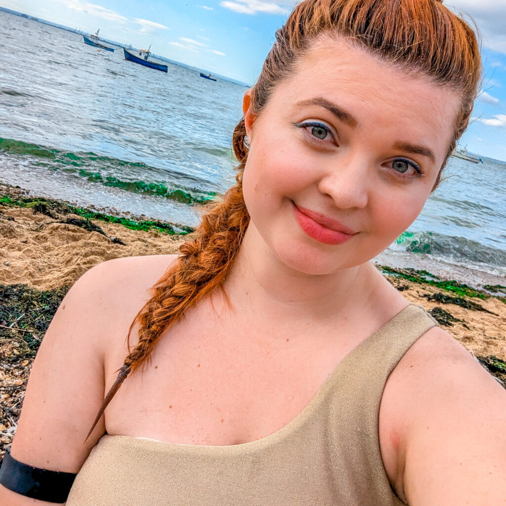 a selfie of Jenni with her hair in a braid she is at the beach wearing a gold swimsuit and her black visible armband