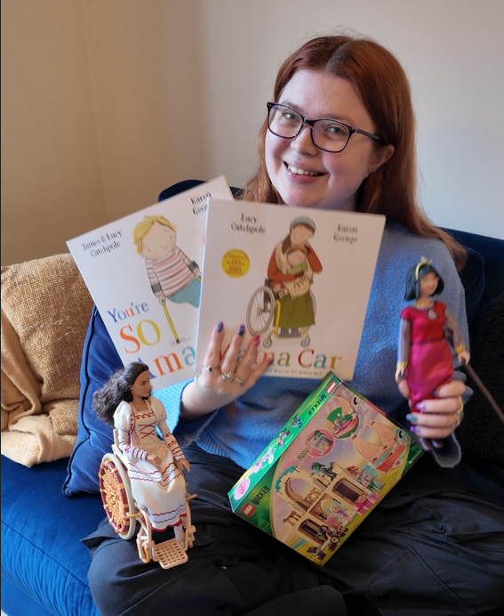 Jenni sitting on her sofa holding a range of inclusive toys with positive disability representation inclusing a nessarose doll, wicked lego set, dahlia doll and books with disability representation