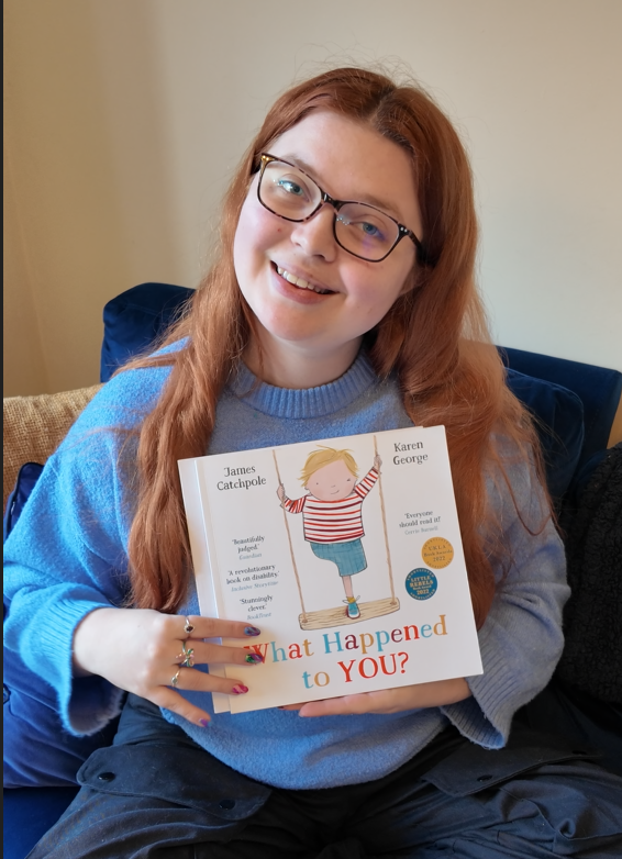 jenni holding a book called what happened to you featuring a young boy on the cover standing on a swing. her has a limb difference is missing 1 leg