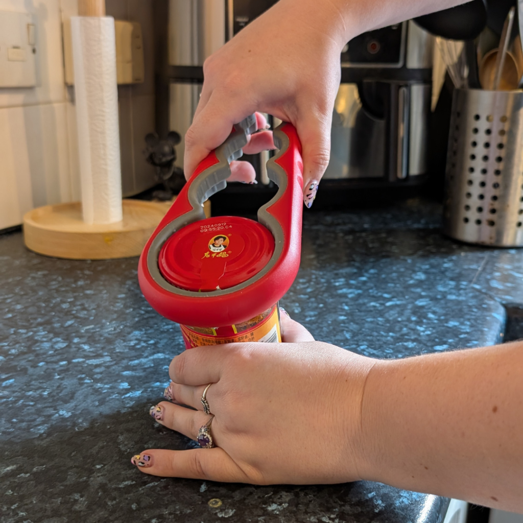 jenni holds a jar in one hand and the red bottle and jar opener tool in the other, it has several different sized round sections to open different sized jars