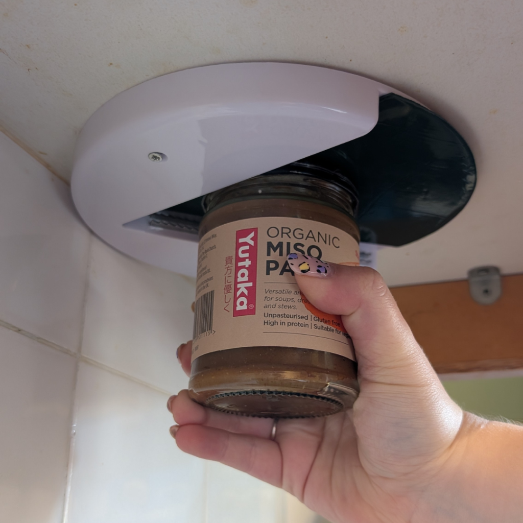 one handed jar opener. attached to the underside of a cupboard is a round white tool with a v shaped black sticky section and metal teeth to hold a jar. Jenni holds a jar in one hand with the lid on the black section to open the jar one handed