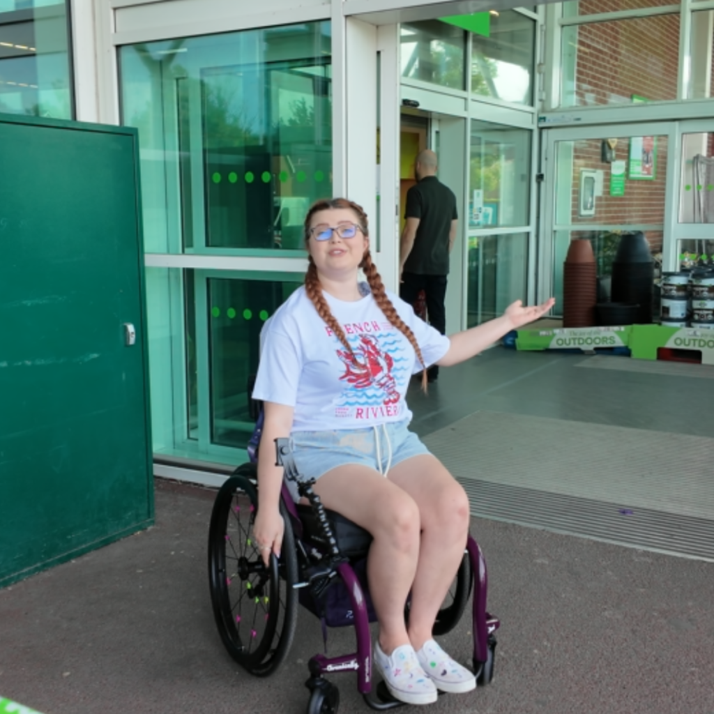 Jenni in a white t-shirt and shorts with two braids indcating towards an automatic door at an asda supermarket. she is sat in her manual wheelchair