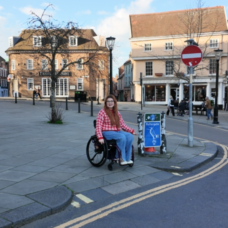 Jenni sitting in her manual wheelchair about to go don one of the original curb cuts in the UK in Norwich