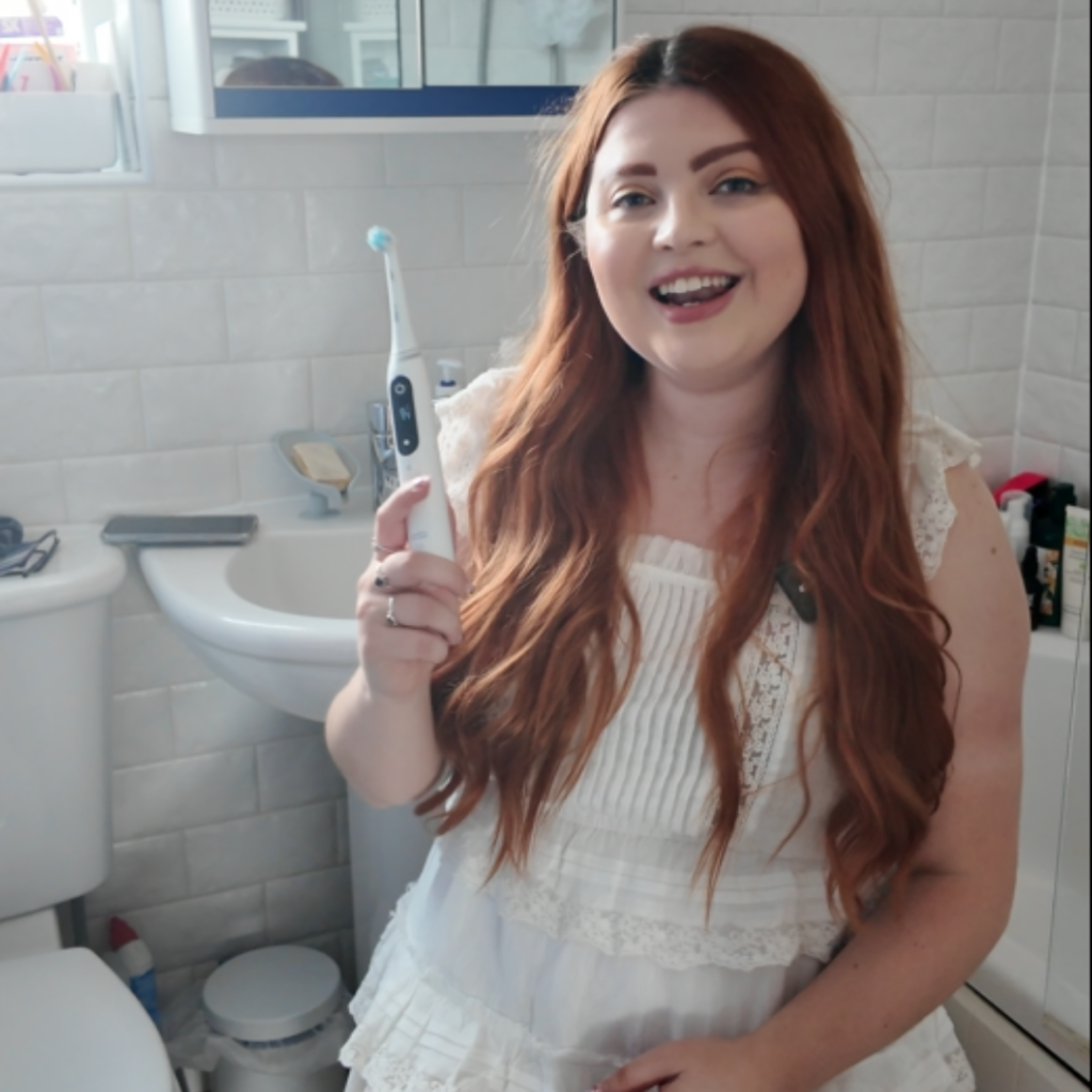 Jenni sat on a stool in he bathroom holding an electric toothbrush. She is wearing a white dress
