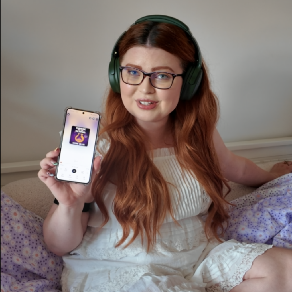 sat on her bed wearing a white dress with her big green headphones on showing her phone to the camera she is listening to the audiobook of sunrise on the reaping