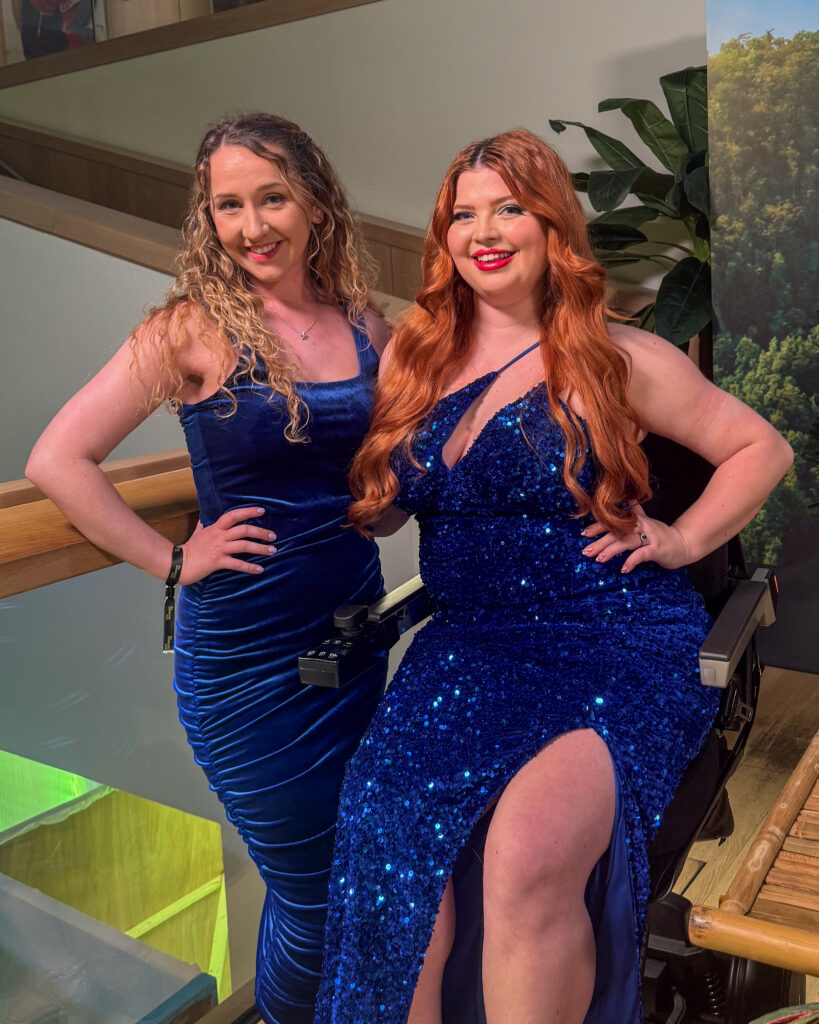 a photo of Jenni and her support worker Beth in sparkly blue dresses. Jenni is sat in her XSTO M4 fully raised up and is almost the same height as Beth who is 5'6 and wearing a small heel.