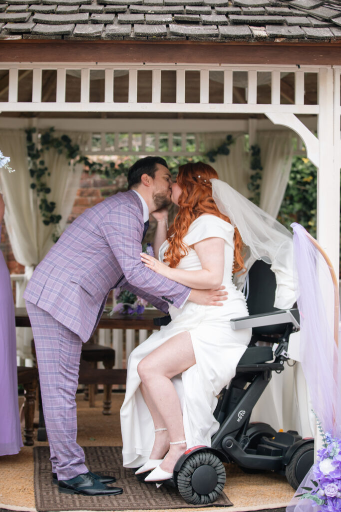 A side on image of Jenni & Ian sharing their first kiss as husband and wife on their wedding day. Jenni is sat raised up in her XSTO M4 powerchair.
