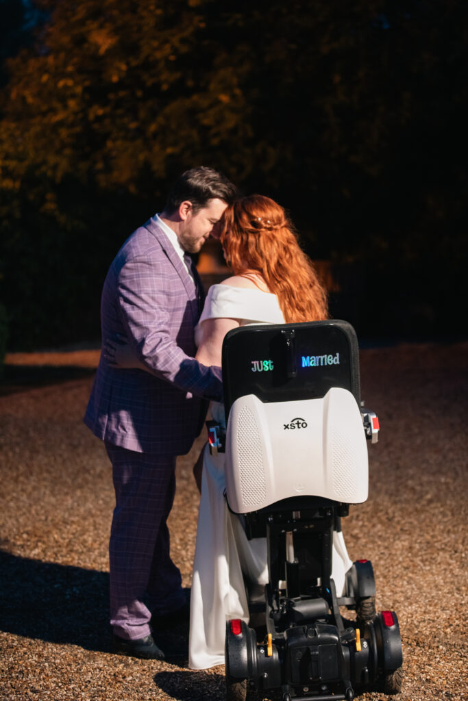 Jenni and Ian with their foreheads together on the evening of their wedding. The back of Jennis XSTO M4 powerchair has a holographic vinyl on the back which says Just Married