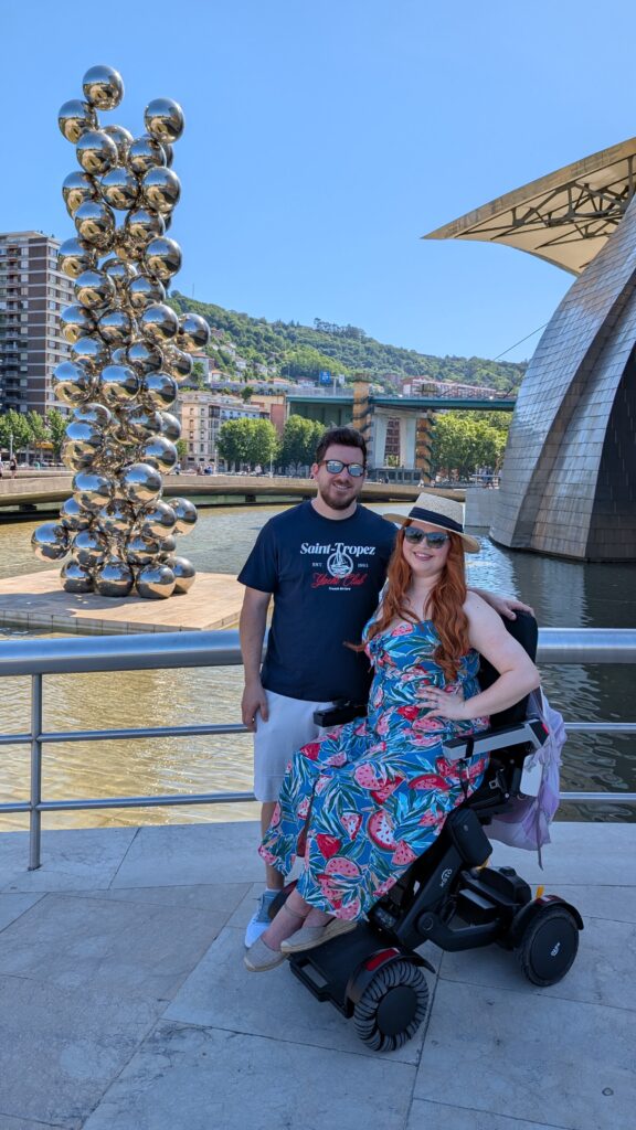 A photo of Jenni & Ian on honeymoon in Bilbao, Spain. They are in front of a statue made of huge silver balls which reflects the light. Jenni is sat side on in her XSTO M4 powerchair and Ian has his arm around the back of her chair