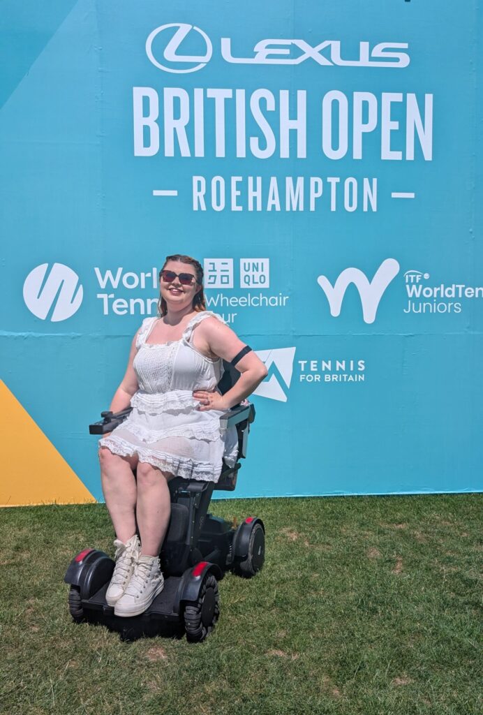jenni wears a white outfit and sits in her XSTO M4 powerchair in front of a turquoise sign for the british open tennis championship